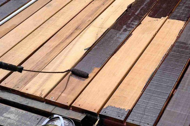 Deck painting professional applying dark protective coating to wooden deck boards with a roller